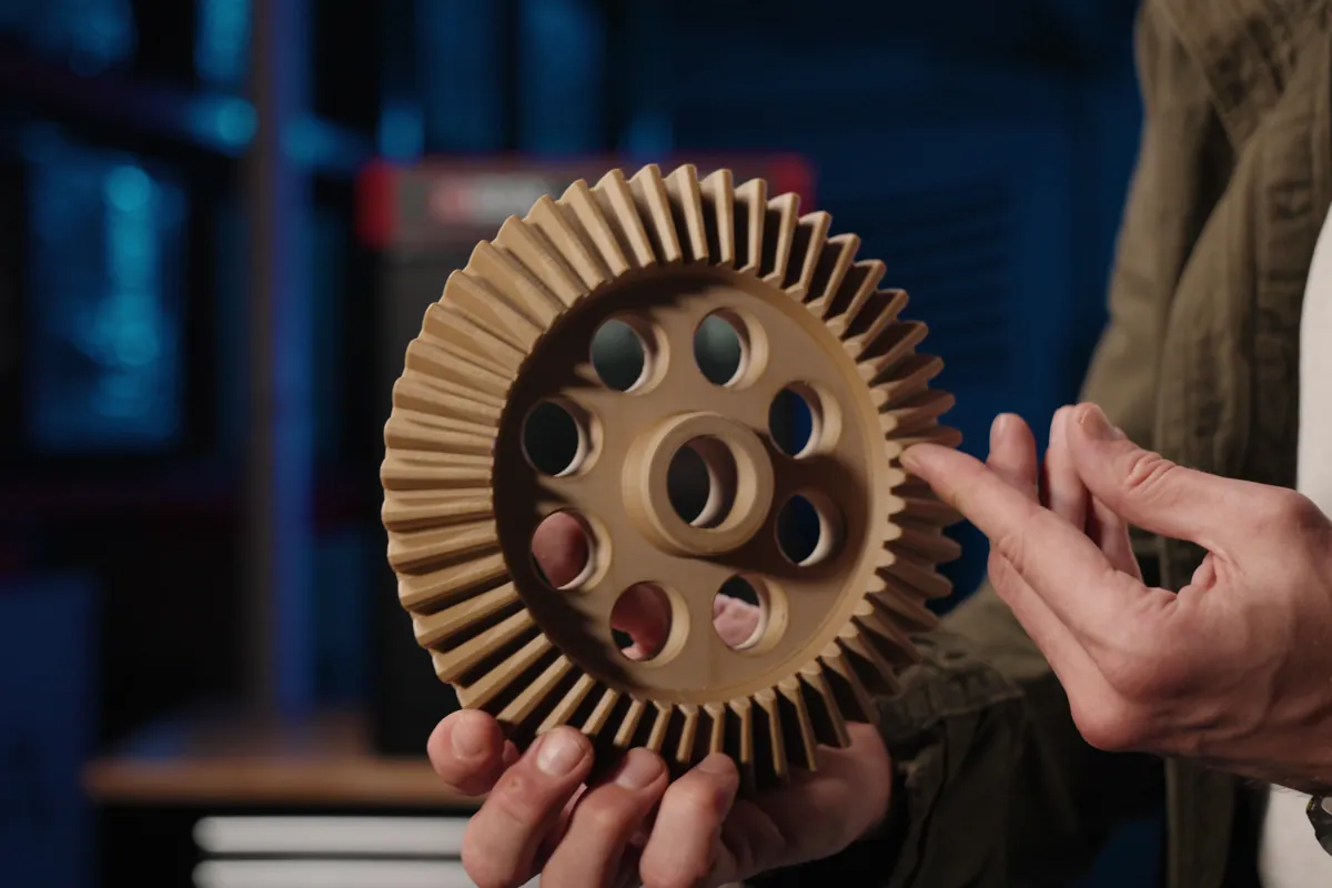 Ingranaggio conico in PEEK caricato vetro stampato in 3D FDM, tenuto in mano davanti a una stampante industriale